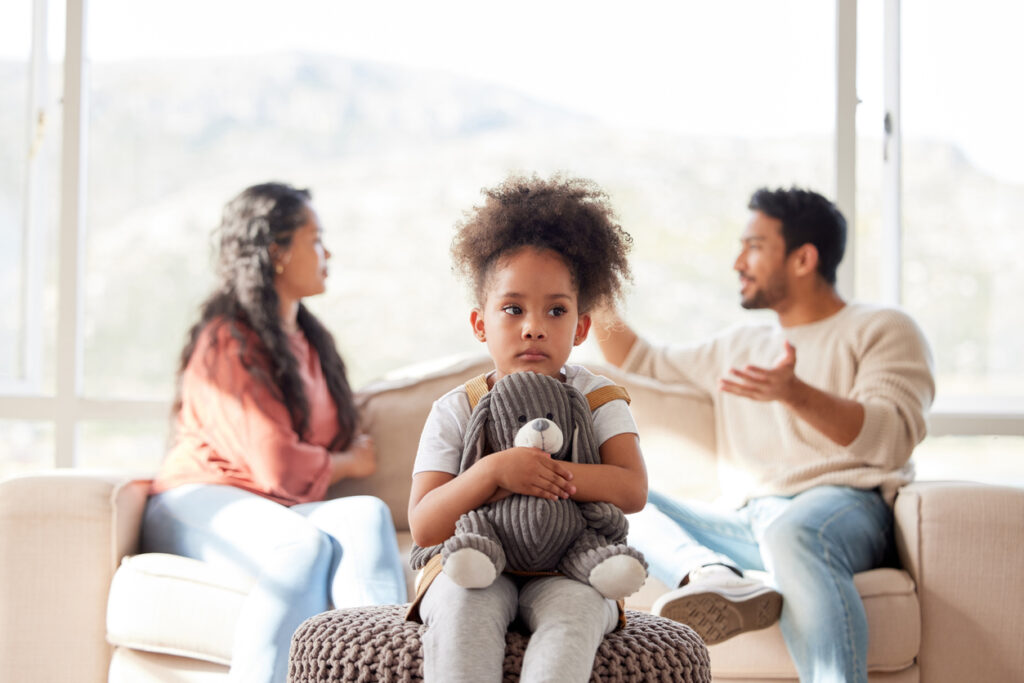 An upset little girl squeezing her teddy bear while looking sad and depressed while her parents argue in the background. Thinking about her parents breaking up or getting divorced is causing stress